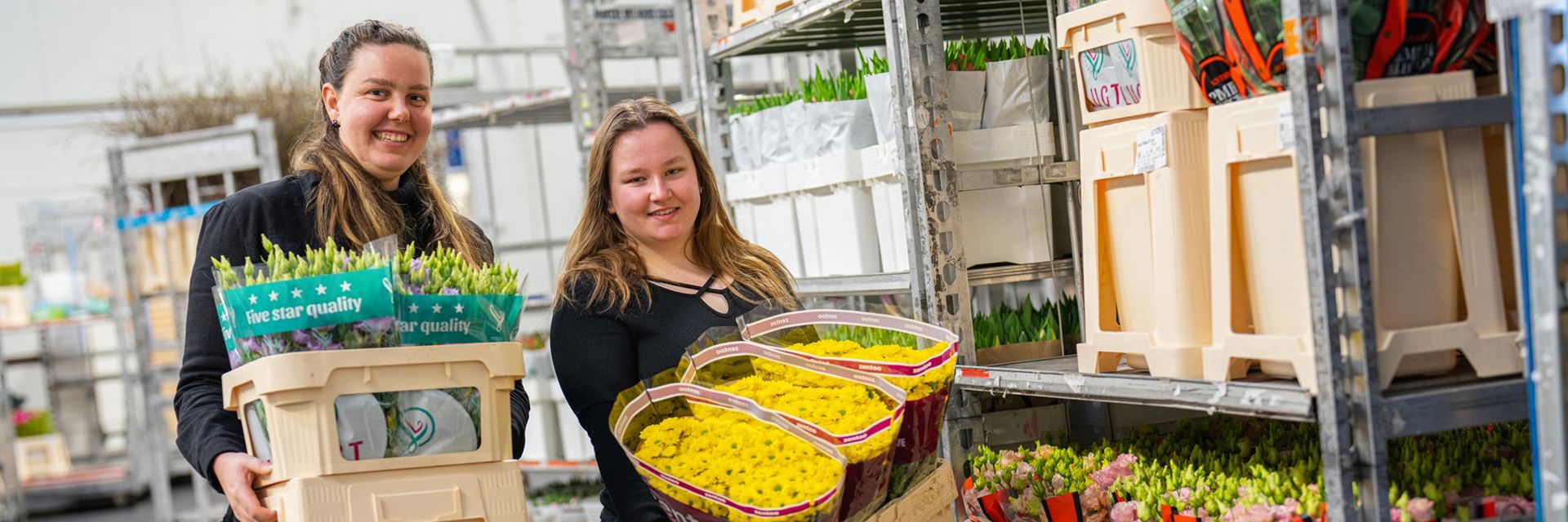 Twee meisjes met een bak bloemen in hun handen, in een bloemenmagazijn